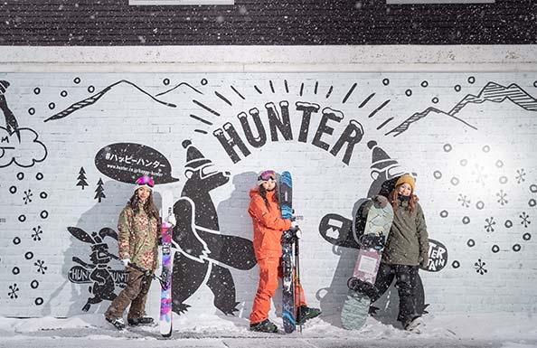 お洒落な壁の前でスノーボードを持つ女性３人組