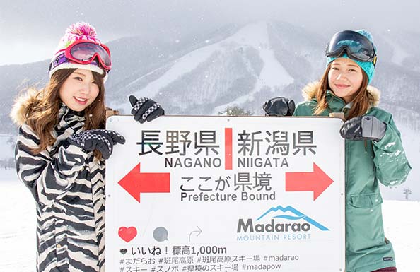 県境の看板の前でポーズを取る女性スノーボーダー２人組